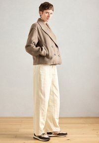 Personne en manteau en laine beige à double boutonnage, pantalon large crème et baskets marron se tient de profil sur un sol en bois contre un mur gris uni.