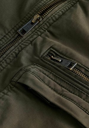 Olive green fabric jacket featuring black zippers, multiple pockets, and stitched detailing, showcasing a smooth texture and functional design.