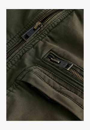 Olive green fabric jacket featuring black zippers, multiple pockets, and stitched detailing, showcasing a smooth texture and functional design.