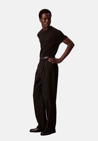 Zwarte t-shirt met korte mouwen, gecombineerd met hoge zwarte broeken en een zwarte riem. De schoenen zijn zwart en gepolijst.