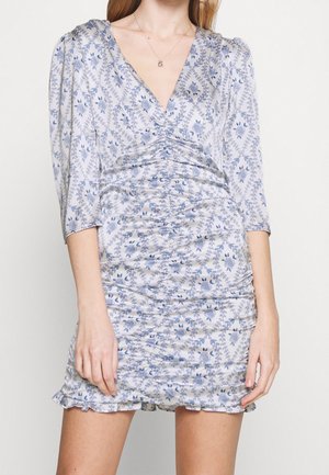 Vrouw draagt een geplooide, lichtblauwe bloemenjurk met driekwartmouwen en een V-hals, staand tegen een eenvoudige achtergrond.