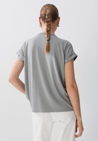 Femme aux cheveux tressés portant des boucles d'oreilles argentées, une chemise grise à manches courtes ample et un pantalon blanc, vue de dos sur un fond blanc.
