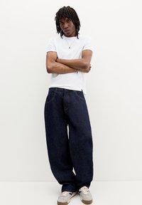 Tricou alb cu mânecă scurtă, blugi dark blue, foarte lejeri, și adidași gri. Detaliile notabile includ cusături vizibile și o siluetă relaxată.