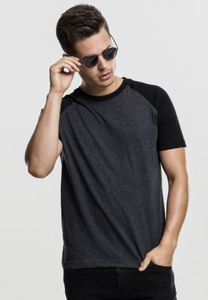 Schwarzes und dunkelgraues T-Shirt mit kurzen Ärmeln und Raglan-Design. Modell trägt Sonnenbrille, Hand auf dem Shirt platziert. Glatte Textur.