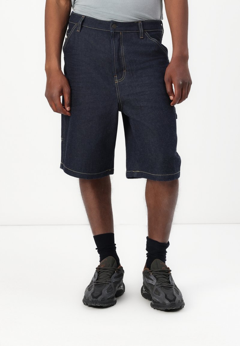 Denimshorts i mørk vask med gule sømmer, med klassisk passform, sidelommer og rett kant. Båret med svarte joggesko og sokker.