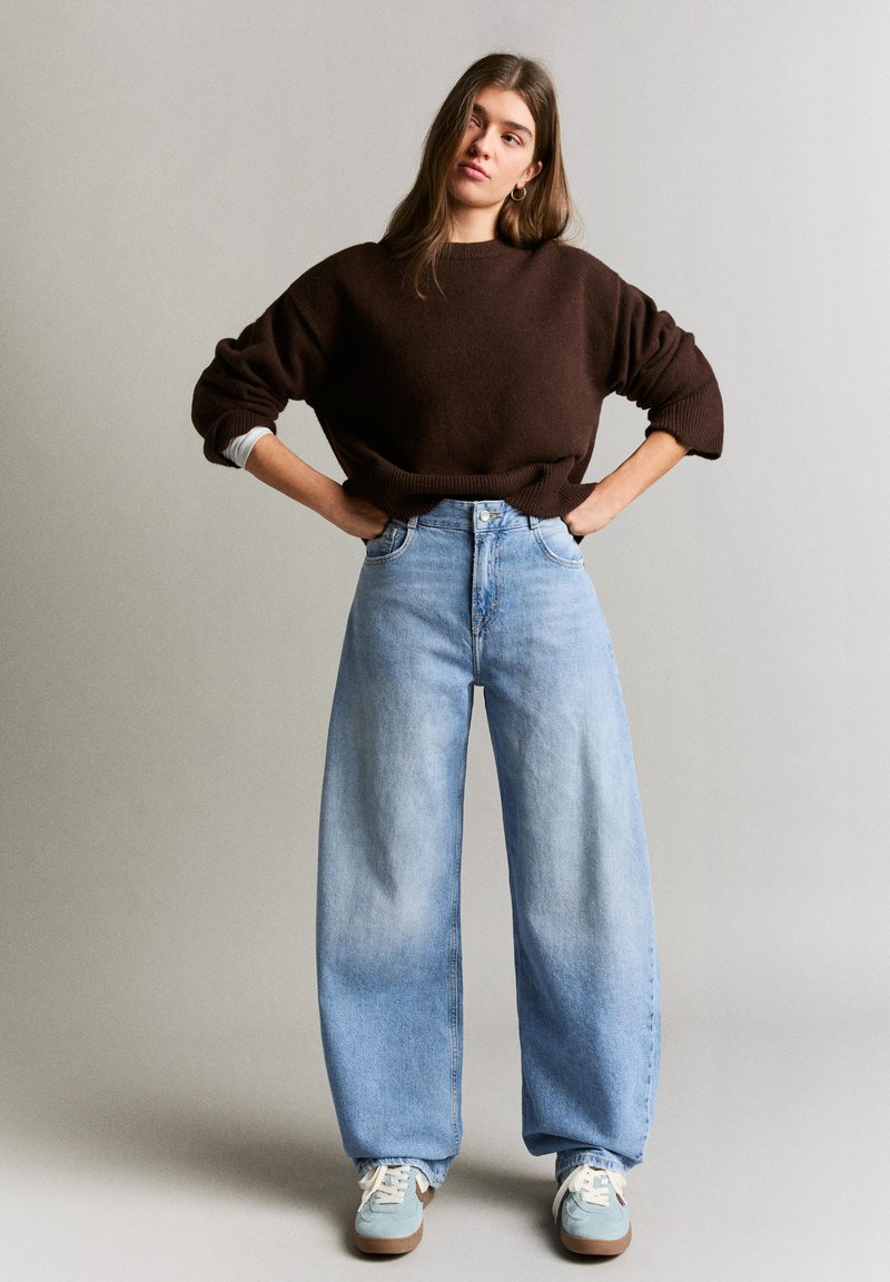 Giovane donna in piedi con le mani sui fianchi, indossa un maglione marrone scuro, jeans a gamba larga color azzurro chiaro e sneakers azzurro chiaro.