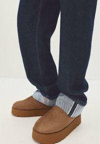 Donkerblauwe jeans omgeslagen bij de enkels, gecombineerd met bruine suède instapschoenen met dikke rubberen zolen.