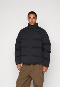 Jachetă puffer neagră cu guler înalt, prevăzută cu închidere cu fermoar și buzunare frontale. Design texturat, capitonat, cu un accent subtil al logo-ului.