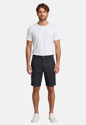 Mann steht und trägt ein schlichtes weißes T-Shirt mit kurzen Ärmeln, schwarze knielange Shorts und weiße Sneakers auf weißem Hintergrund.