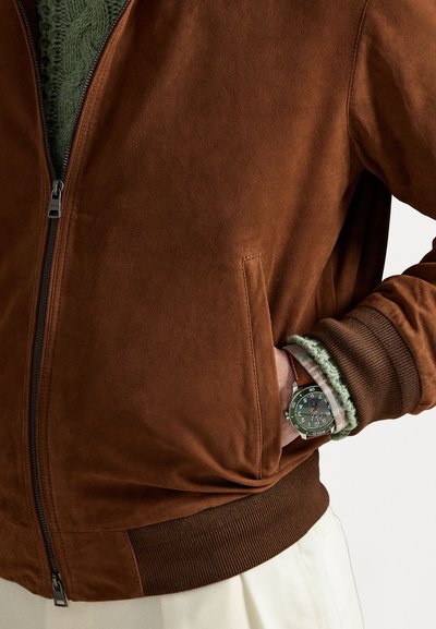 Braune Wildleder-Bomberjacke mit gerippten Bündchen und Saum. Grüner Strickunterzieher, der am Kragen sichtbar ist. Silberne Armbanduhr mit grünem Ziffernblatt am Handgelenk.