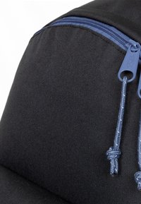 Zaino nero con una superficie testurizzata, caratterizzato da una cerniera blu e dettagli in corda coordinati. La forma è arrotondata, enfatizzando il suo design compatto.