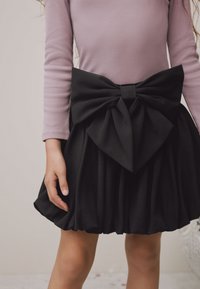 Gonna nera plissettata con un grande fiocco in vita, indossata sopra una maglia a maniche lunghe color lilla chiaro. Tessuto morbido con una texture liscia.