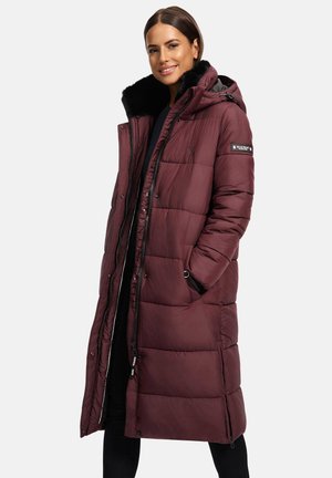 Woman wearing a long maroon padded coat with black fur-lined hood, hands in pockets, smiling, standing against a plain light background.