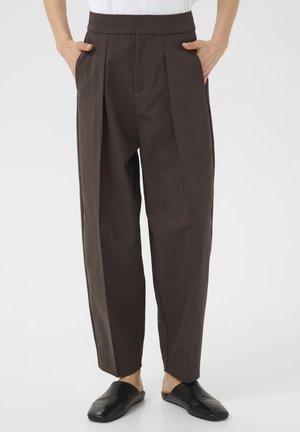 Brune højtaljede bukser med folder, lige pasform og sidelommer, parret med sorte slip-on sko. Glat stofstruktur.