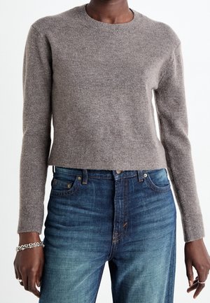 Person iført en taupe langærmet striktrøje og blå højtaljede jeans, med sølvarmbånd og ringe på venstre hånd.