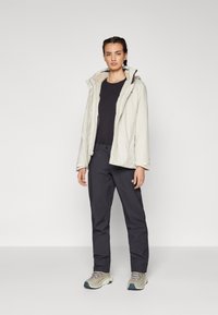 Giacca impermeabile grigio chiaro con colletto foderato in pelliccia, abbinata a pantaloni neri e sneaker chiare con accenti grigi e blu.