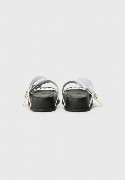 Hvide sandaler med en mat finish, der har to krydsede stropper og en sort sål. Guld hardware detalje på siden. Flad design.