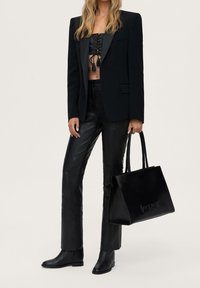 Sac cabas en cuir noir avec logo embossé, associé à un blazer noir cintré, un haut noir ajusté et un pantalon en cuir noir orné.