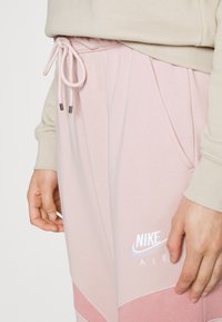 Osoba v světle růžových teplácích Nike Air s šňůrkou v pase a béžové mikině, ruce uvolněně po stranách.