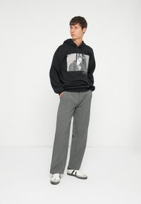 Felpa nera con una stampa grafica, abbinata a pantaloni grigi su misura e sneakers bianche con strisce nere. Design minimalista, look casual.