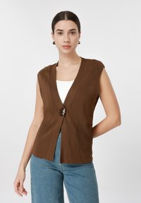 Γυναίκα με σκουρόχρωμα μαλλιά που φοράει καφέ αμάνικη ζακέτα πάνω από λευκή μπλούζα και μπλε τζιν, στέκεται μπροστά σε λευκό, απλό φόντο.