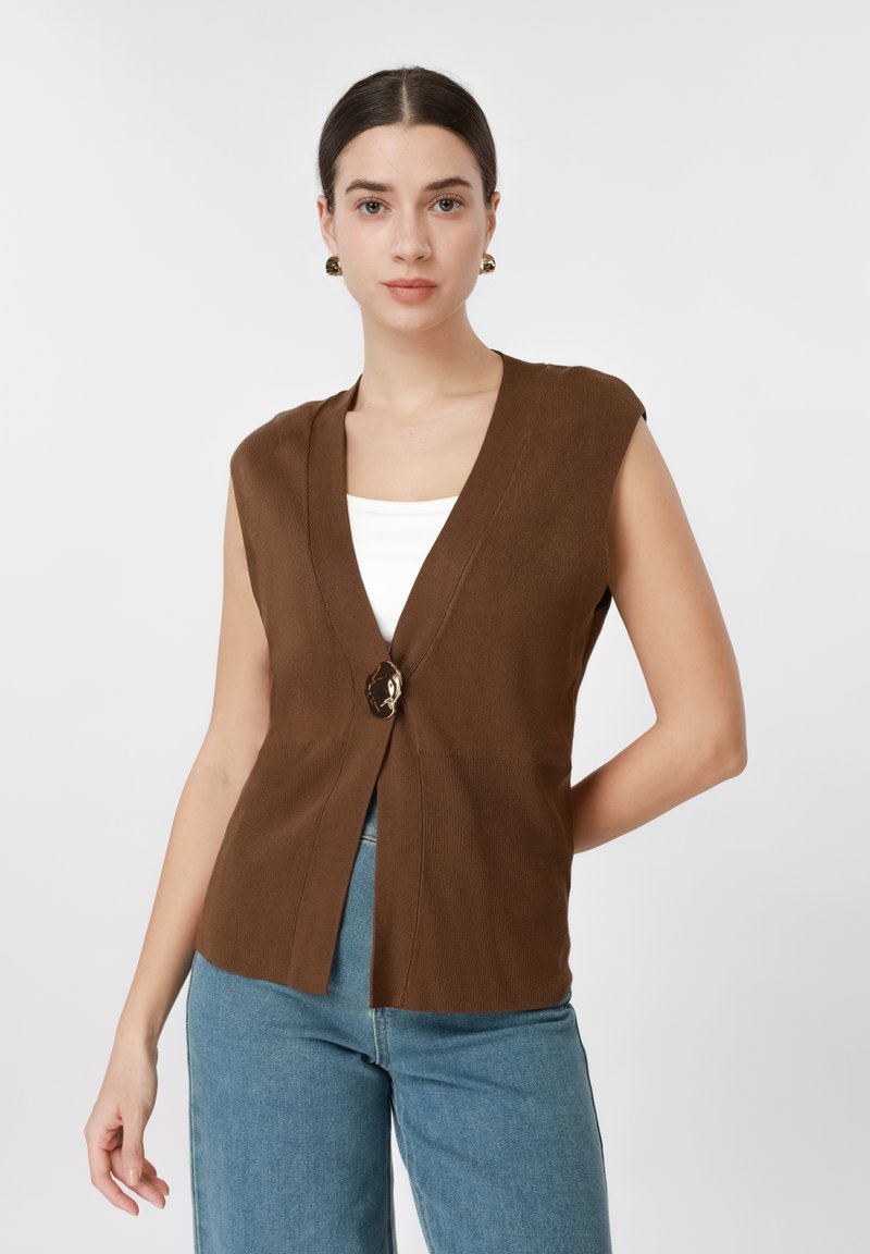 Γυναίκα με σκουρόχρωμα μαλλιά που φοράει καφέ αμάνικη ζακέτα πάνω από λευκή μπλούζα και μπλε τζιν, στέκεται μπροστά σε λευκό, απλό φόντο.