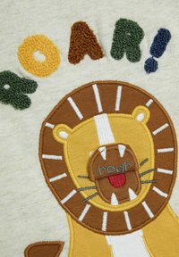 Vyšívaný design lva na světlém materiálu s textem "ROAR!". Mezi jeho vlastnosti patří hnědé a žluté odstíny, texturovaná hříva a hravé detaily na obličeji.