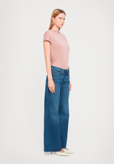 Haut rose clair à manches courtes associé à un jean en denim bleu taille large. Modèle portant des chaussures blanches à enfiler et de grandes boucles d'oreilles en métal doré.
