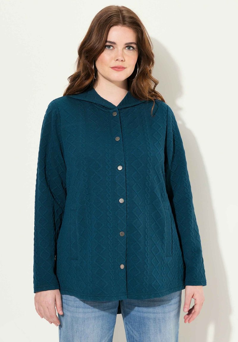 Teal quiltad jacka med huva, prydd med ett texturerat diamantmönster, metallknäppningar och två sidofickor. Långa ärmar och avslappnad passform.