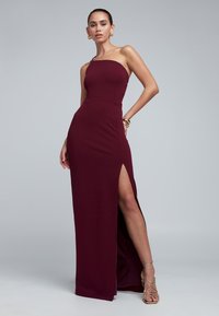 Burgundska, oprijeta obleka z enim ramenom in visokim razporkom, gladka tkanina in minimalističen dizajn. Okrašena z zlatim nakitom.