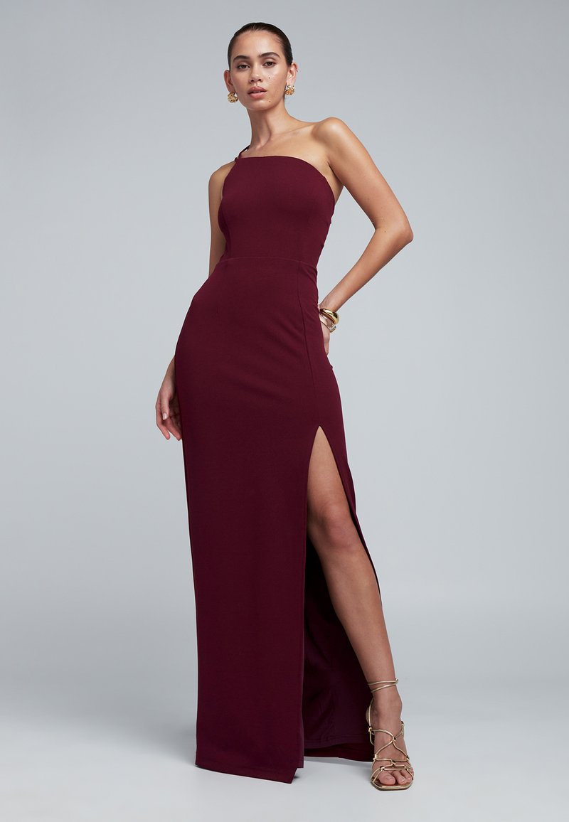 Burgundska, oprijeta obleka z enim ramenom in visokim razporkom, gladka tkanina in minimalističen dizajn. Okrašena z zlatim nakitom.