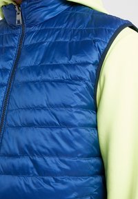 Niebieska kamizelka puchowa o błyszczącej, pikowanej teksturze, z wysokim kołnierzem i zapięciem na zamek, noszona na jasnozielonej polarowej bluzie z kapturem.