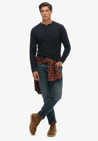 Camisa henley de manga larga en color navy con tres botones, combinada con jeans azul y botas de cordones marrones. Una camisa de franela de cuadros en rojo y negro atada a la cintura.
