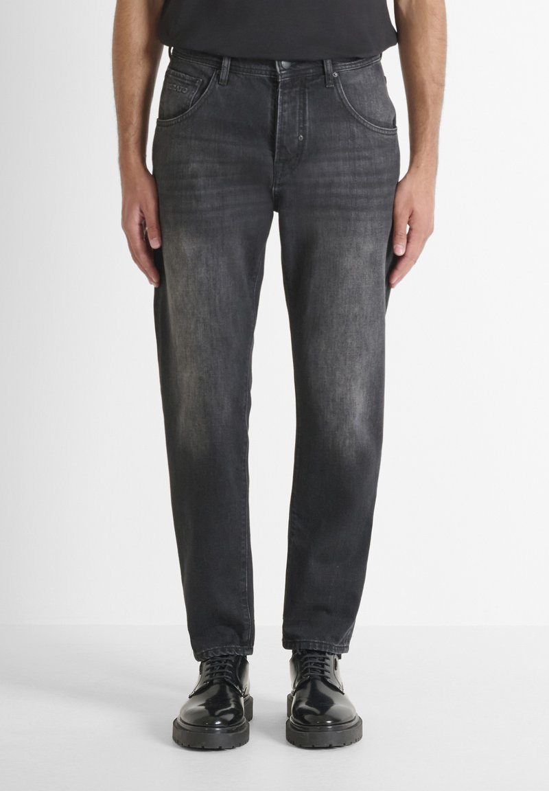 Jeans de mezclilla negra con un acabado ligeramente desgastado, diseño de cinco bolsillos y corte recto. Combinados con zapatos negros brillantes con cordones.