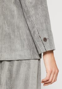 Manica di un blazer in velluto a coste grigio con righe verticali testurizzate, dotato di una chiusura a bottone marrone sul polsino, abbinato a pantaloni coordinati.