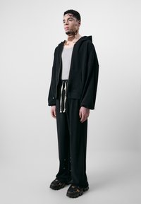 Schwarze Zip-Hoodie mit Fronttasche, graues Tanktop, lockere schwarze Sweatpants mit Kordelzug und klobige schwarze Schuhe mit braunen Akzenten.