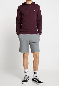 Bordó mikina s kapucí a klokaní kapsou s žlutým logem, doplněná o šedé šortky a černé tenisky s bílými detaily a pruhované ponožky.