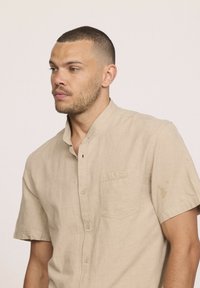Chemise à manches courtes en lin beige. Présente une poche plaquée et une patte de boutonnage. Tissu texturé avec une coupe décontractée.