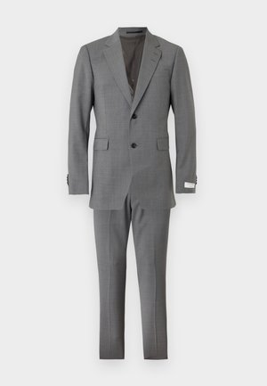 Ung mand stående, iført en grå slim-fit jakkesæt med matchende bukser, lyseblå skjorte og sorte læder loafers, med hvid baggrund.