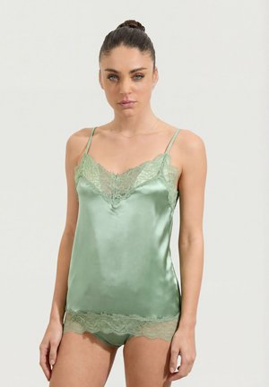 Woman wearing light green silk camisole and matching lace-trimmed underwear, standing against plain light background.