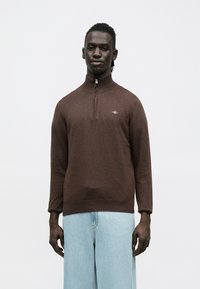 Brūns džemperis ar augstu apkakli un puszipa aizdari. Izgatavots no mīksta auduma, ar ribotām aprocēm un smalku logotipa detaļu.