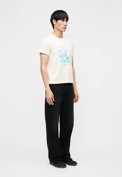 Jeune homme debout portant un T-shirt blanc avec un motif de chaton bleu, un pantalon noir à jambes larges et des chaussures noires sur un fond uni.
