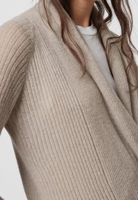 Cárdigan de punto acanalado beige con un cuello de chal y textura suave. Cuenta con un frente drapeado, que muestra la calidad ligera de la tela.