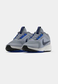 Grå idrottsskor med blå accenter, mesh-oversida och en vit sula. Har en rundad tå och framträdande logotyp på sidan och plösen.