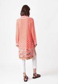 Garšs, plūstošs oranžs un sarkans kimono ar ģeometriskiem rakstiem, ar apkakli un garām piedurknēm, valkāts virs baltām īsām biksēm.