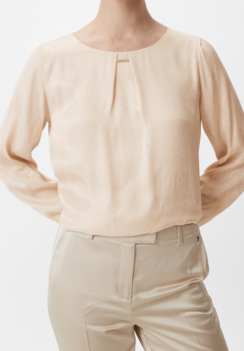 Kvinde iført beige satin bluse med lange ærmer og lille frontfold samt matchende beige bukser med høj talje.