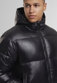 Juodas pūkinis sportinis švarkas blizgiu paviršiumi, su užtrauktuku, gobtuvu ir horizontaliais siūlais. Šiltas, izoliuotas dizainas, tinkamas šaltam orui.