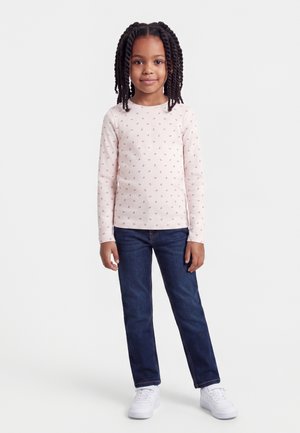 Jeune fille aux cheveux tressés portant un t-shirt rose clair à manches longues avec de petits motifs, un jean foncé et des baskets blanches, debout sur un fond blanc.