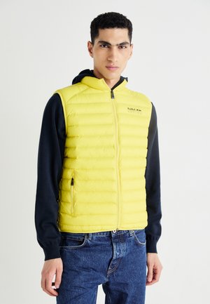 Chaleco acolchado amarillo con diseño de cuadros horizontales, cremallera frontal y bolsillos laterales. Combinado con una sudadera con capucha azul marino de manga larga y jeans de mezclilla.