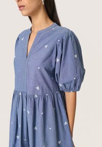 Modra chambray obleka s napihnjenimi rokavi, V-izrezom in belim cvetličnim vezenjem. Večplastna zasnova z udobnim krojem in mehko teksturo.